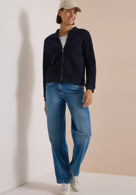 Femme en veste bleue, jean et casquette appuyée décontractée contre un mur clair.