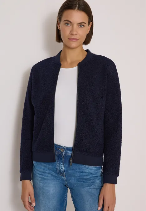 Femme aux cheveux bruns courts porte une veste polaire bleu foncé, un t-shirt blanc et un jean bleu.