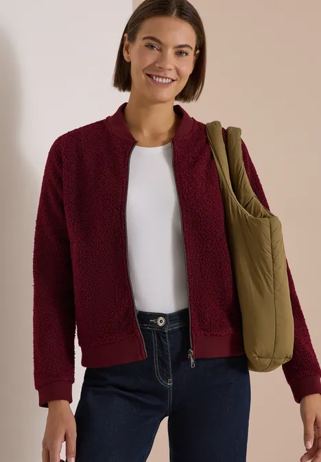 Femme souriante en cardigan bordeaux, chemise blanche, jean foncé et grand sac olive porté à l'épaule.