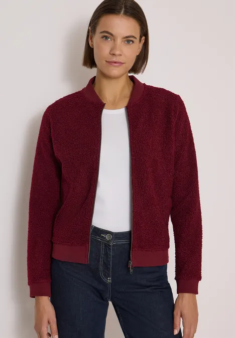 Femme en veste teddy bordeaux, t-shirt blanc et jean foncé sur fond clair.