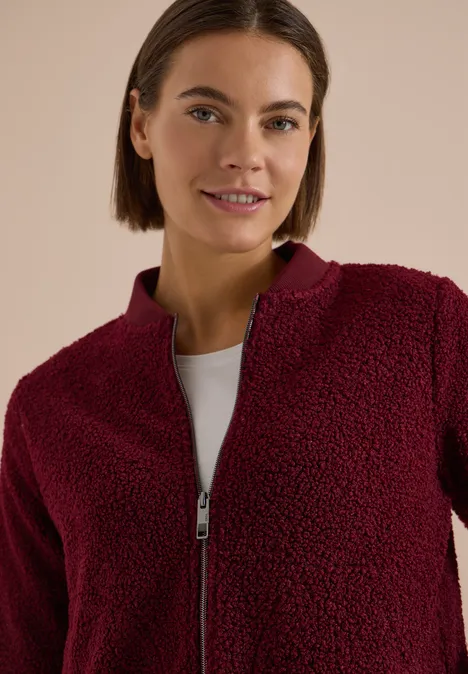 Femme brune aux cheveux courts porte un pull zippé en polaire bordeaux et un t-shirt blanc sur fond neutre.
