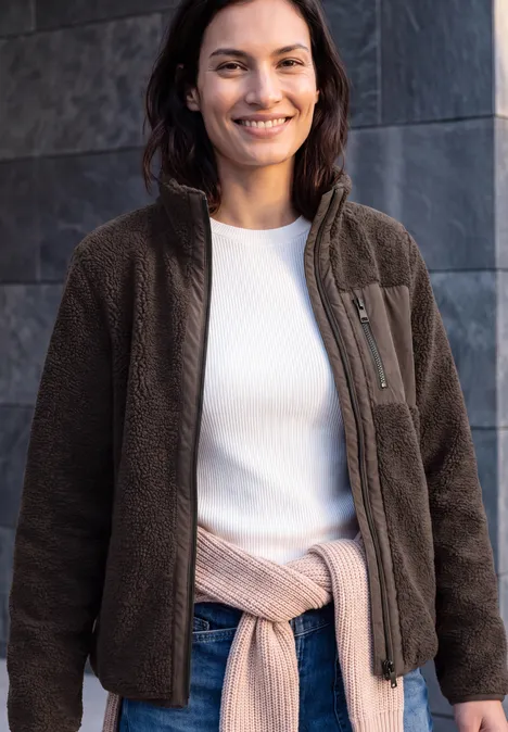 Eine lächelnde Frau trägt eine braune Fleecejacke, ein weißes Shirt und hat einen Pullover um die Hüfte gebunden.