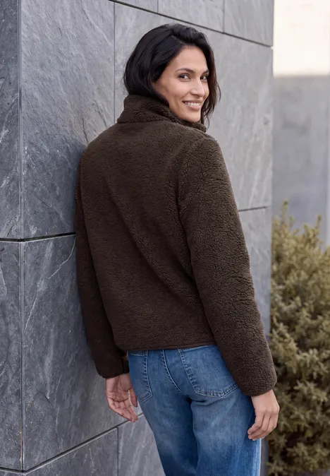 Eine Frau steht seitlich vor einer grauen Steinwand, trägt braune Fleecejacke und blaue Jeans, blickt über die Schulter.