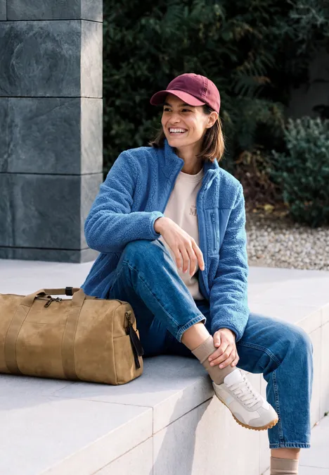 Eine Frau mit roter Mütze und blauer Fleecejacke sitzt lächelnd mit Tasche auf einer Steinbank.