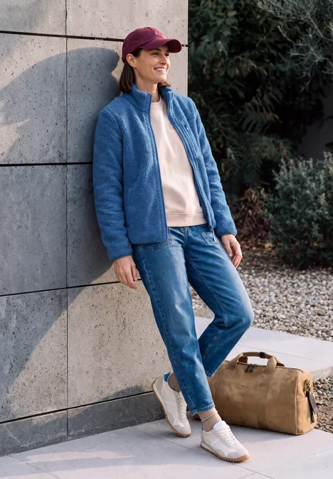 Eine junge Frau lehnt an einer Wand, trägt Jeans, blaue Jacke, pinke Mütze, lächelt, Reisetasche neben ihr.