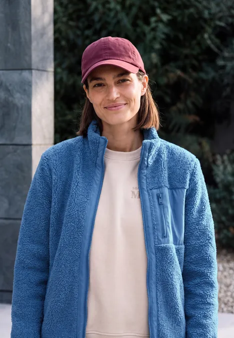 Frau mit roter Kappe und blauer Fleecejacke lächelt vor grünem Hintergrund.