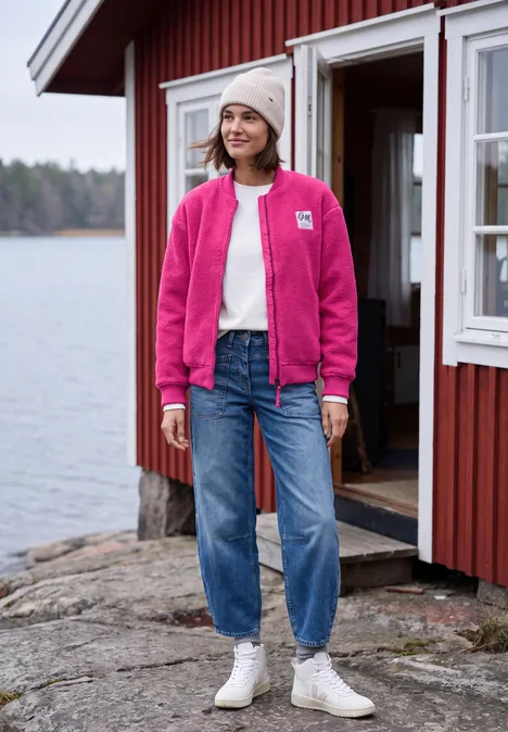 Frau mit pinker Jacke, Jeans und Mütze steht vor roter Hütte am See.
