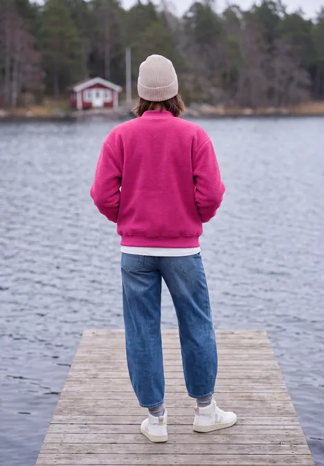 Person in pinkem Pullover und weißer Mütze steht auf Steg, blickt über ruhigen See mit Waldhaus.