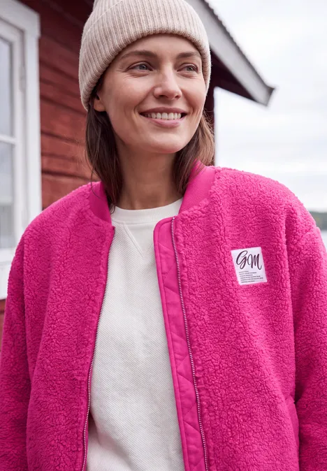 Frau in pinkem Teddy-Fleece-Jacke und Beanie lächelt vor rotem Haus.
