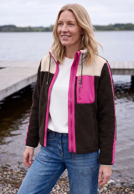 Teddy-Jacke mit Softshelldetails truffle brown Frau in braun-weißer Jacke mit pinken Akzenten, Jeans und weißem Pullover, lächelt am Ufer eines Sees.