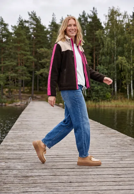 Teddy-Jacke mit Softshelldetails truffle brown Frau in bunter Jacke und Jeans spaziert lächelnd auf einem Steg am See vor bewaldetem Hintergrund.