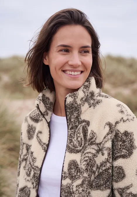 Teddyjasje met opstaande kraag en patroon teddy vanilla white Vrouw lacht op het strand, draagt een gemêleerde fleecejas, duinen op de achtergrond.