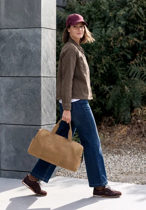 Frau mit roter Kappe, brauner Jacke und blauer Jeans trägt eine große hellbraune Tasche beim Spaziergang.