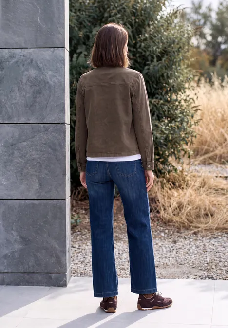 Eine Person steht mit dem Rücken zur Kamera in Outdoor-Umgebung, trägt braune Jacke, Jeans und braune Schuhe.