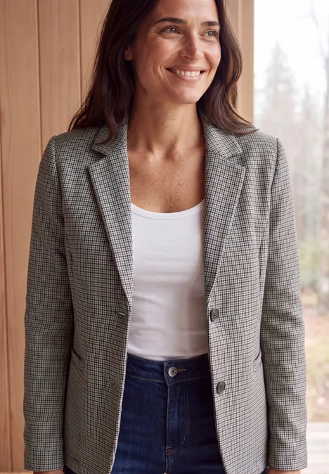 Frau lächelt in grauem Glencheck-Blazer, weißem Top und Jeans vor hellem Holzhintergrund.