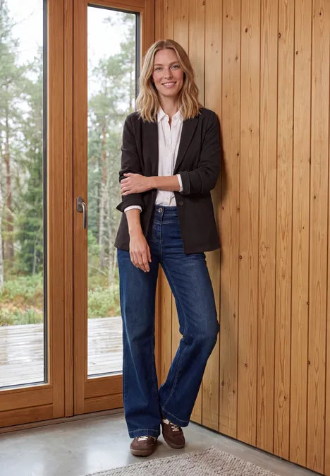Een glimlachende vrouw in jeans en blazer staat voor een houten wand naast grote glazen deuren.