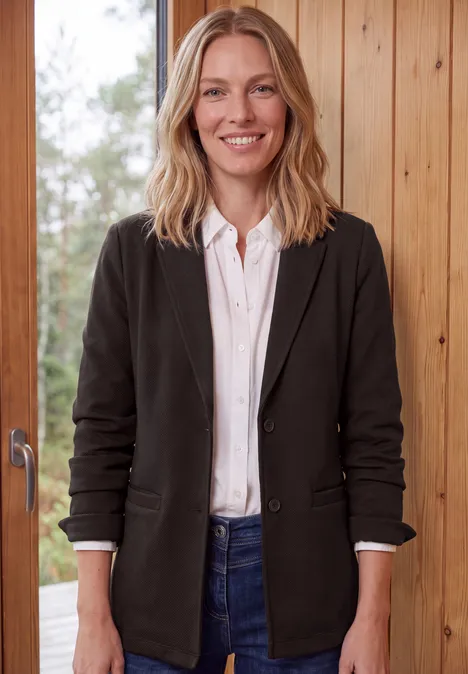 Een glimlachende blondine draagt een zwarte blazer, wit overhemd en jeans voor een houten achtergrond.