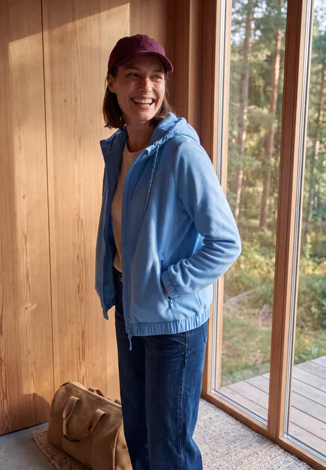 Lächelnde Frau in blauer Jacke und Jeans steht mit Tasche in hellem Raum vor Fenstern im Wald.