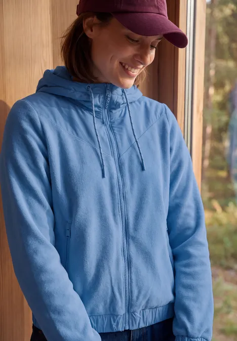 Junge Frau im blauen Kapuzen-Sweatshirt und Cap lächelt stehend vor einem Fenster mit Waldkulisse.