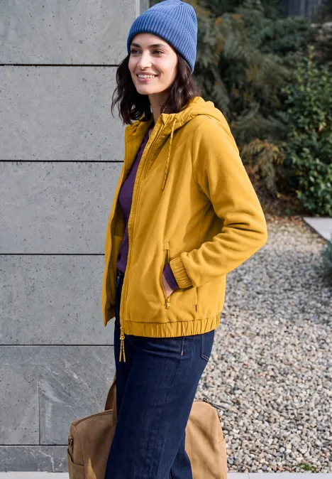 Fleecejas met capuchon en softshell-details golden honey Eine Frau in gelber Jacke, blauer Mütze und Jeans steht lächelnd vor einer grauen Wand und hält eine Tasche.