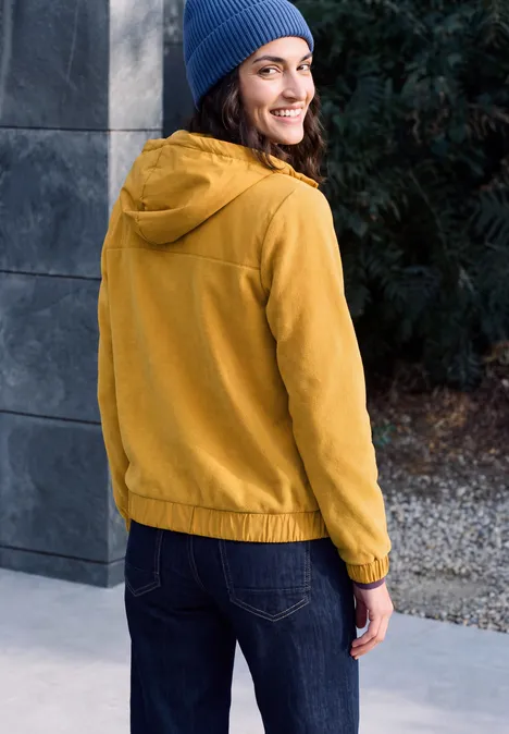 Fleecejas met capuchon en softshell-details golden honey Eine Frau steht im Freien, trägt eine gelbe Jacke und eine blaue Mütze und blickt lächelnd über die Schulter.