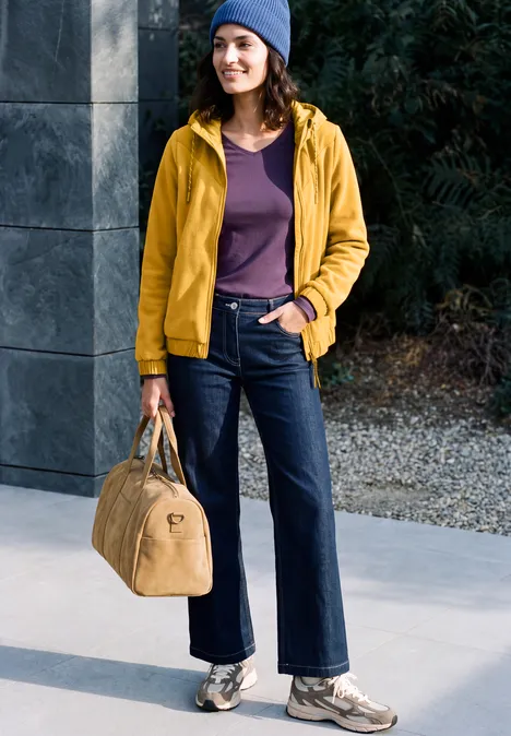 Fleecejas met capuchon en softshell-details golden honey Frau in gelber Jacke, Jeanshose und Mütze mit Reisetasche in der Hand, posiert lächelnd im Freien.