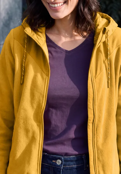 Fleecejas met capuchon en softshell-details golden honey Frau trägt gelbe Kapuzenjacke über lila T-Shirt, nur Oberkörper und leichtes Lächeln sichtbar.
