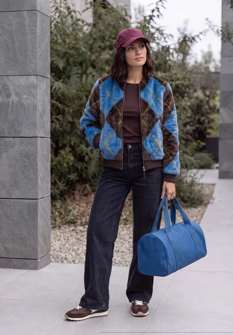 Junge Frau im karierten Mantel, Jeans, Hut und blaue Tasche steht draußen vor einem modernen Gebäude.