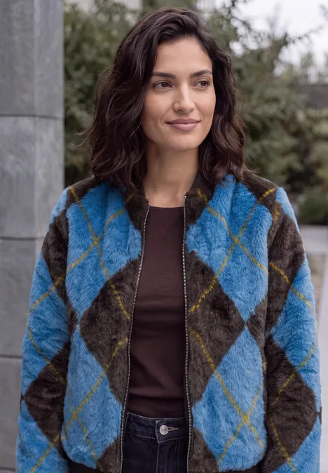 Eine Frau trägt eine türkis-graue Rauten-Fleecejacke und ein braunes Shirt, steht draußen und lächelt leicht.