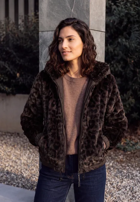 Fake Fur Jacke mit Softshell-Kapuze truffle brown Eine Frau mit lockigem Haar trägt eine kuschelige schwarze Jacke und steht auf Kies vor einer Betonwand.
