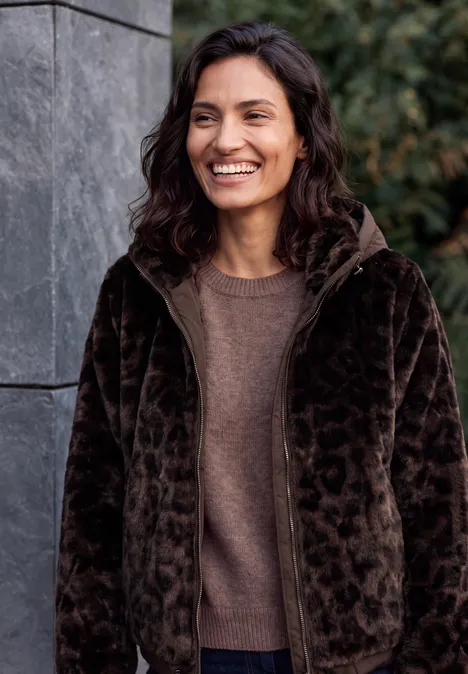 Fake Fur Jacke mit Softshell-Kapuze truffle brown Lächelnde Frau in braunem Pullover und flauschiger, dunkler Jacke vor einer Steinwand mit grünen Pflanzen.