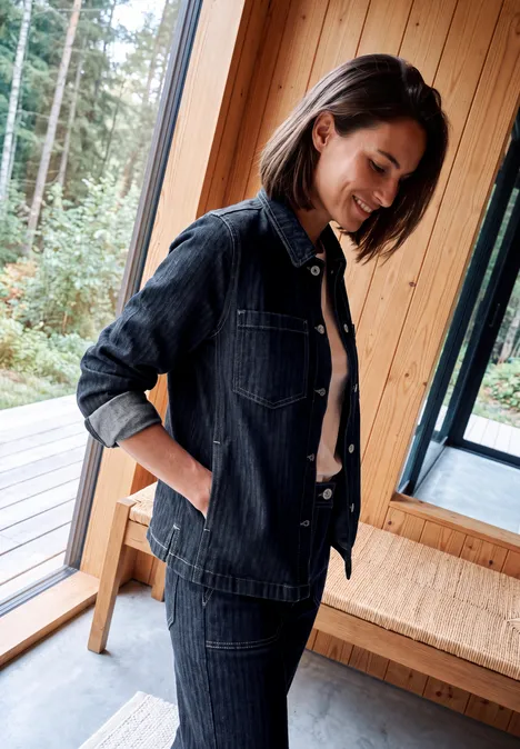 Femme en jean sombre souriant près d’une fenêtre dans une pièce en bois lumineux.