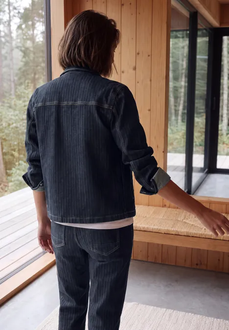 Personne de dos en veste et pantalon en jean dans un intérieur en bois moderne avec grandes fenêtres.