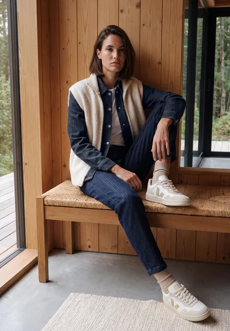 Jeune femme en jean et veste douillette assise détendue sur un banc en bois devant une baie vitrée.