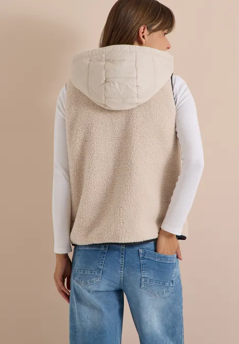 Rückenansicht einer Frau in cremefarbener, wattierter Weste mit Kapuze, weißem Shirt und blauen Jeans.
