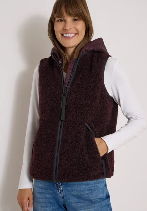 Eine lächelnde Frau trägt ein weißes Langarmshirt, eine dunkelrote Fleece-Weste mit Kapuze und Jeans.