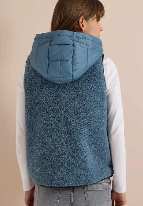 Frau von hinten in jeansblauer Teddyweste mit gesteppter Kapuze und weißem Langarmshirt.