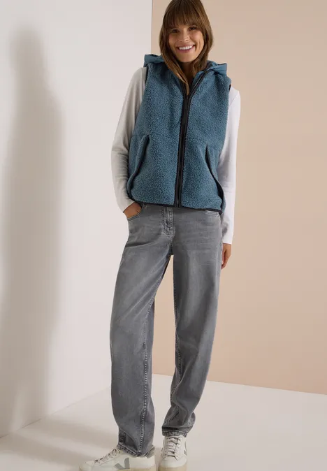 Eine Frau trägt einen grauen Hoodie, eine blaue Fleece-Weste und eine weite Jeans.