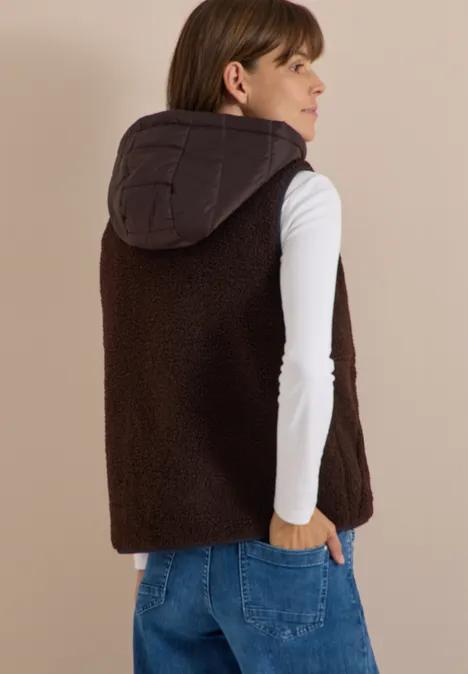 Frau mit weißem Langarmshirt, blauer Jeans und brauner Teddyfell-Weste mit Kapuze, Rücken zur Kamera.