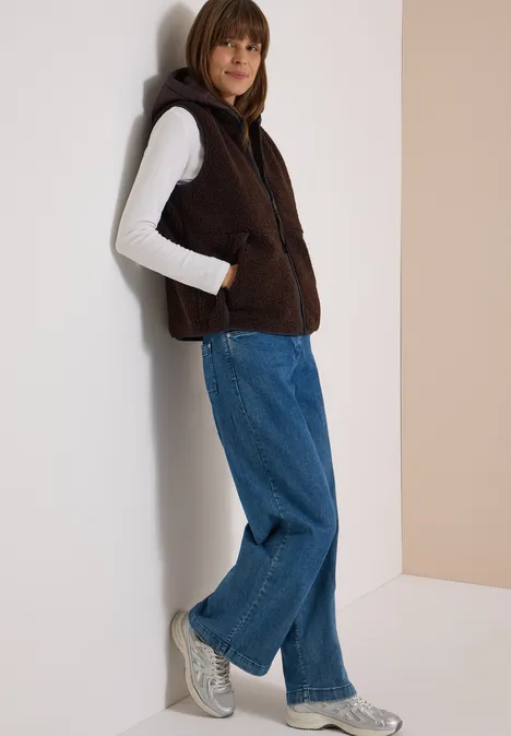 Frau lehnt an Wand, trägt braune Fleece-Weste mit Kapuze, weißen Pullover, weite blaue Jeans und graue Sneaker.
