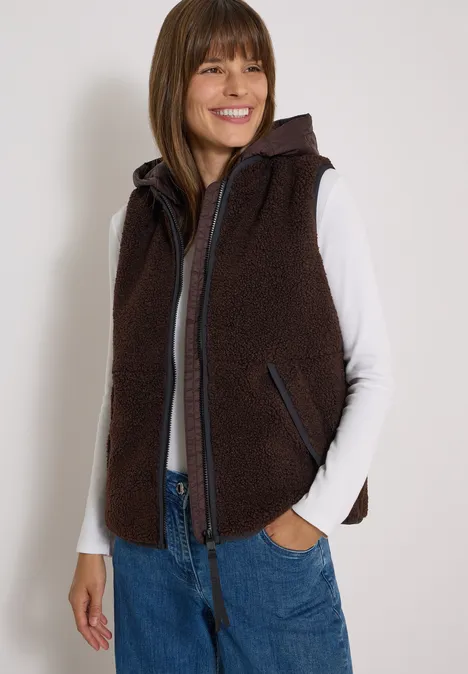 Frau trägt braune Teddy-Fleece-Weste mit Kapuze, Reißverschluss und Eingrifftaschen über weißem Shirt und Jeans.
