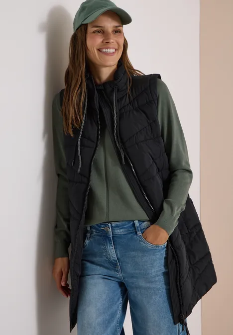 Lange Steppweste mit Stehkragen Black Frau mit grüner Baseballcap, grünem Pullover, blauen Jeans und langer schwarzer Steppweste lächelt.