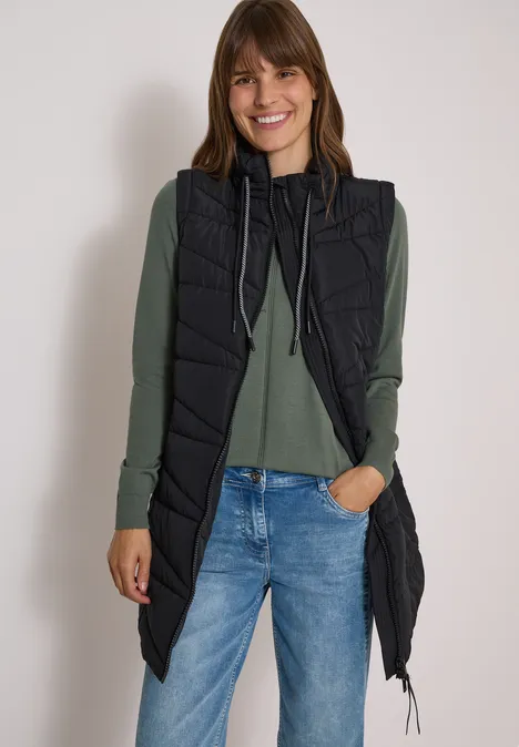 Lange Steppweste mit Stehkragen Black Frau in langer schwarzer Steppweste, grünem Pullover und blauer Jeans, lächelnd vor weißem Hintergrund.