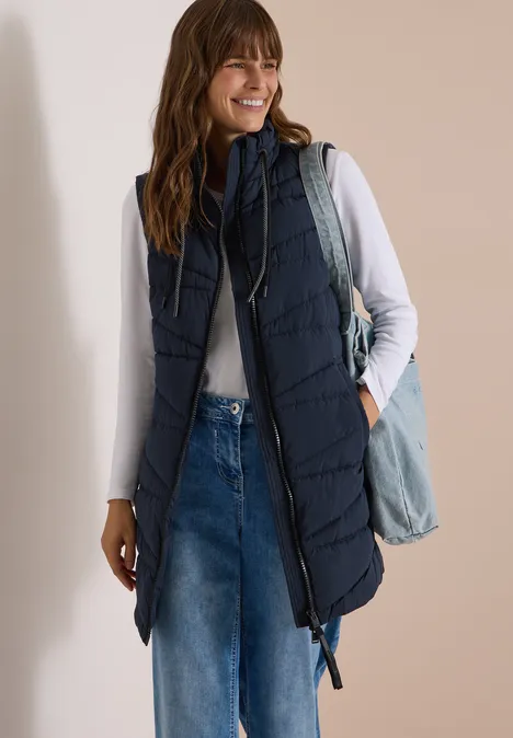 Lange Steppweste mit Stehkragen dark navy blue Frau in weißem Langarmshirt, blauer ärmelloser Steppweste, Jeans und Umhängetasche.