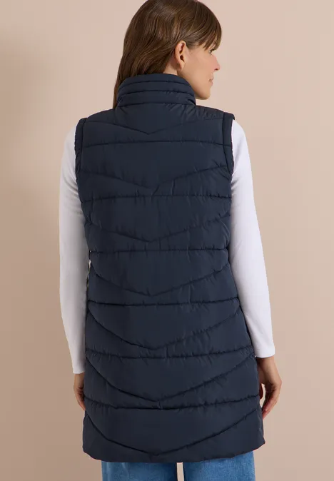 Lange Steppweste mit Stehkragen dark navy blue Frau von hinten in weißem Langarmshirt und navyblauer, knielanger Steppweste vor beigem Hintergrund.