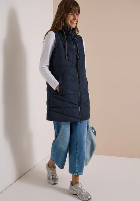 Lange Steppweste mit Stehkragen dark navy blue Frau mit dunkelblauer Steppweste, weißem Shirt, weitem Jeans, silbernen Sneakers und Tasche.