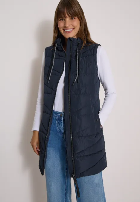 Lange Steppweste mit Stehkragen dark navy blue Frau trägt weiße Bluse, blaue Jeans und lange dunkelblaue Steppweste mit Kapuze und Reißverschluss.