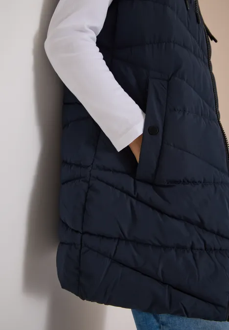 Lange Steppweste mit Stehkragen dark navy blue Seitliche Ansicht einer dunkelblauen Steppweste mit Taschen, dazu weißes Shirt und Jeans.
