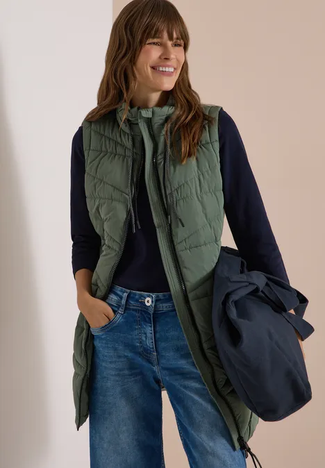 Lange Steppweste mit Stehkragen shadow khaki Frau in grüner, langer, gesteppter Weste über dunklem Pullover und Jeans, hält eine Jacke in der Hand.