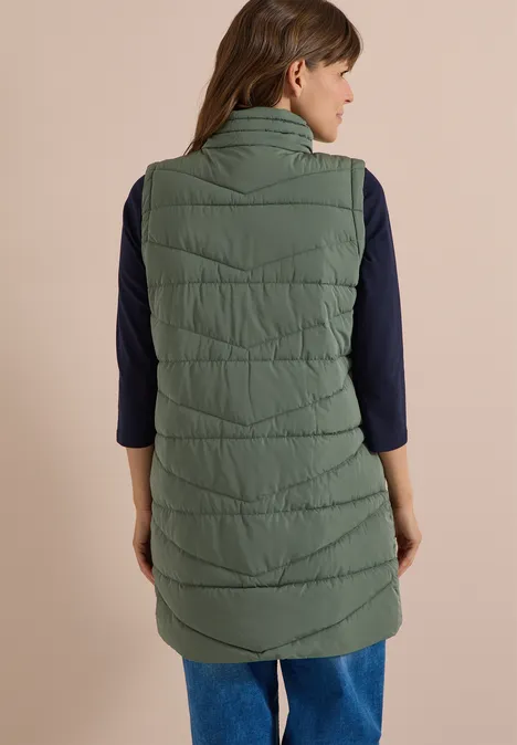 Lange Steppweste mit Stehkragen shadow khaki Frau von hinten in langer olivgrüner Steppweste, dunkelblauem Shirt und Jeans.